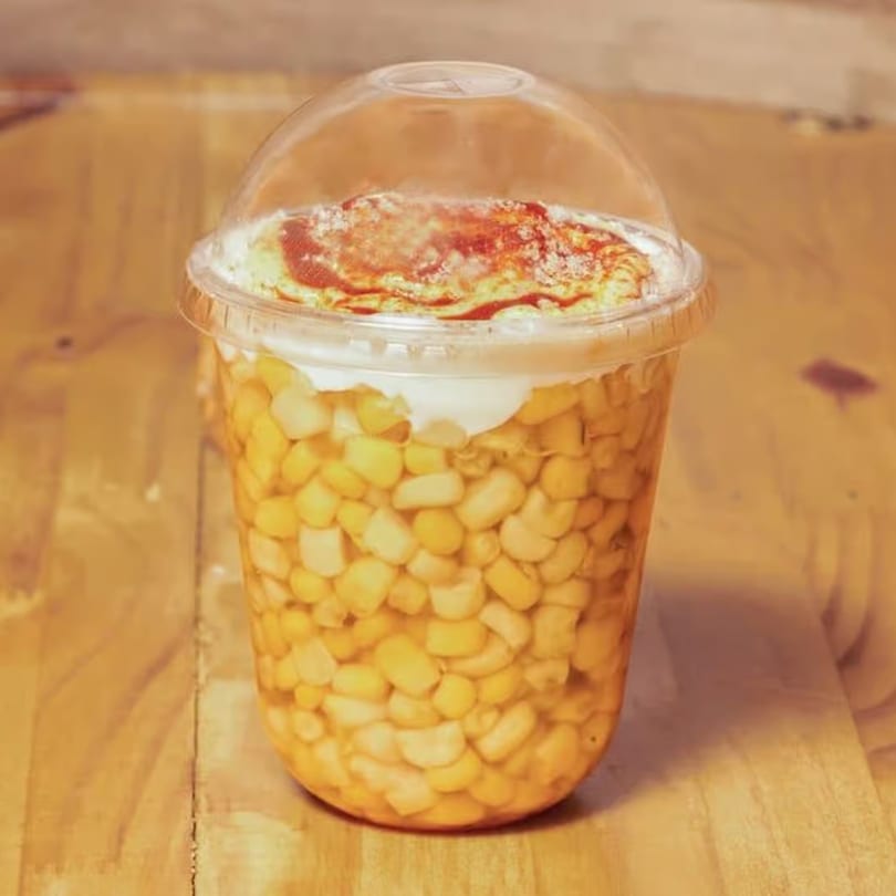 A clear plastic cup filled with corn kernels, topped with white cream and red chili powder.