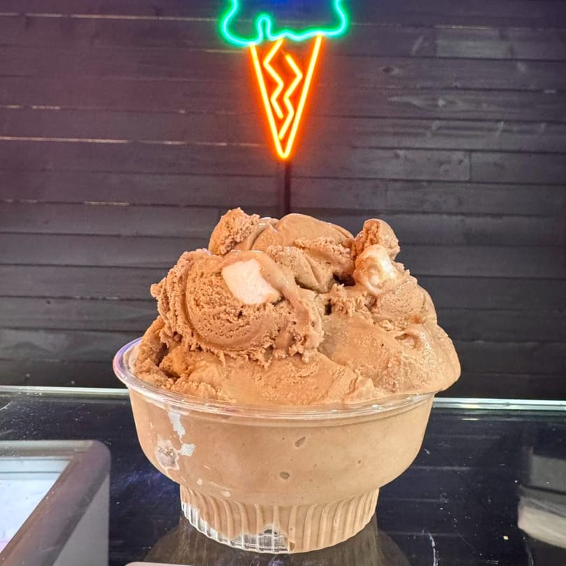 A large scoop of chocolate ice cream in a clear cup under a neon ice cream cone sign.