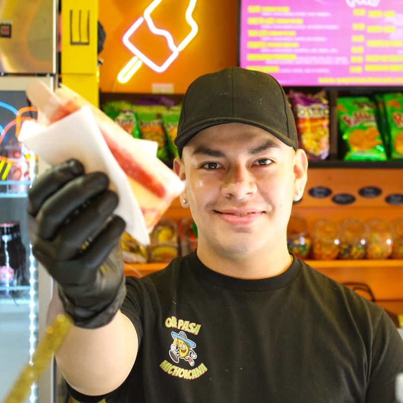 A smiling Que Pasa Michoacana employee in a black cap and shirt offers a colorful paleta.