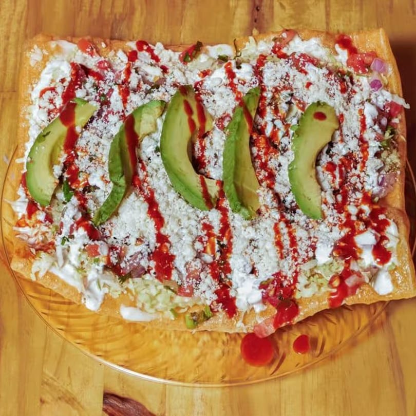 A large rectangular tostada topped with avocado slices, white cheese, red sauce, and crema.