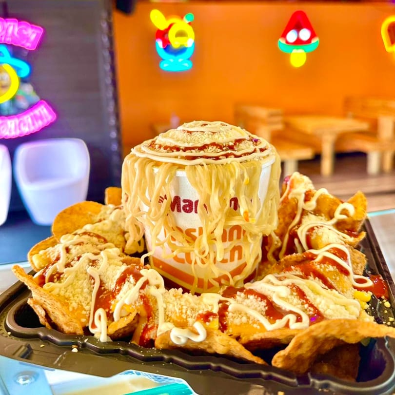 A cup of ramen noodles topped with sauce and cheese, surrounded by tortilla chips with similar toppings.
