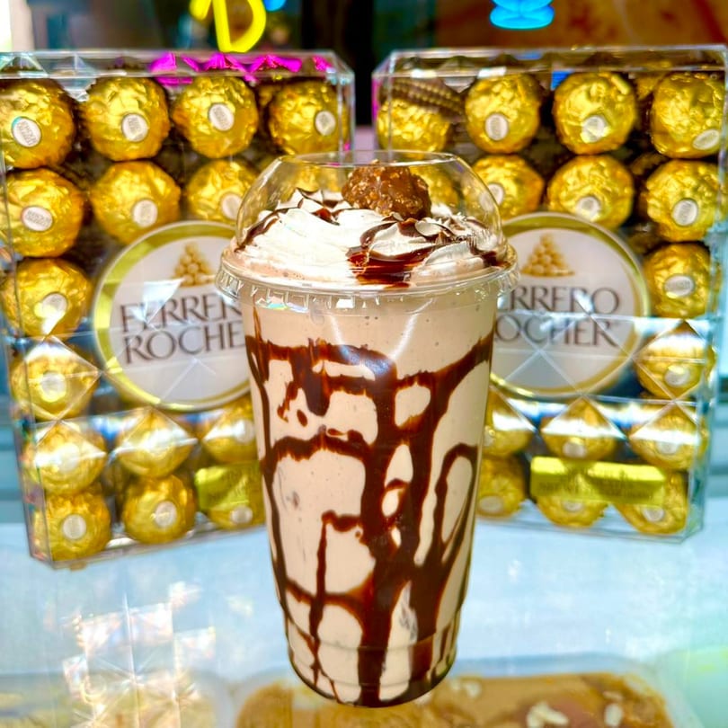 Ferrero Rocher milkshake with whipped cream and chocolate drizzle, surrounded by boxes of Ferrero Rocher chocolates.