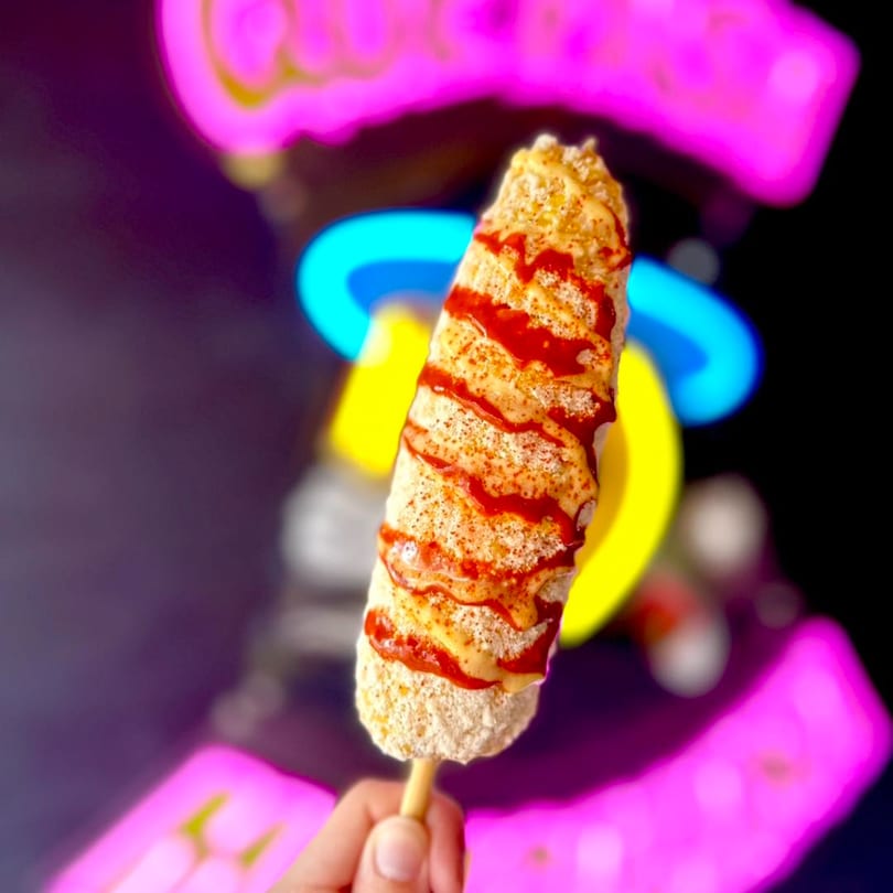 Hand holding an elote, a Mexican street corn, drizzled with sauce and seasoning.