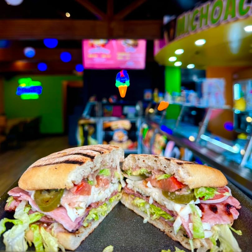 A large, grilled sandwich cut in half, filled with meats, cheese, lettuce, tomatoes, and jalapeños.