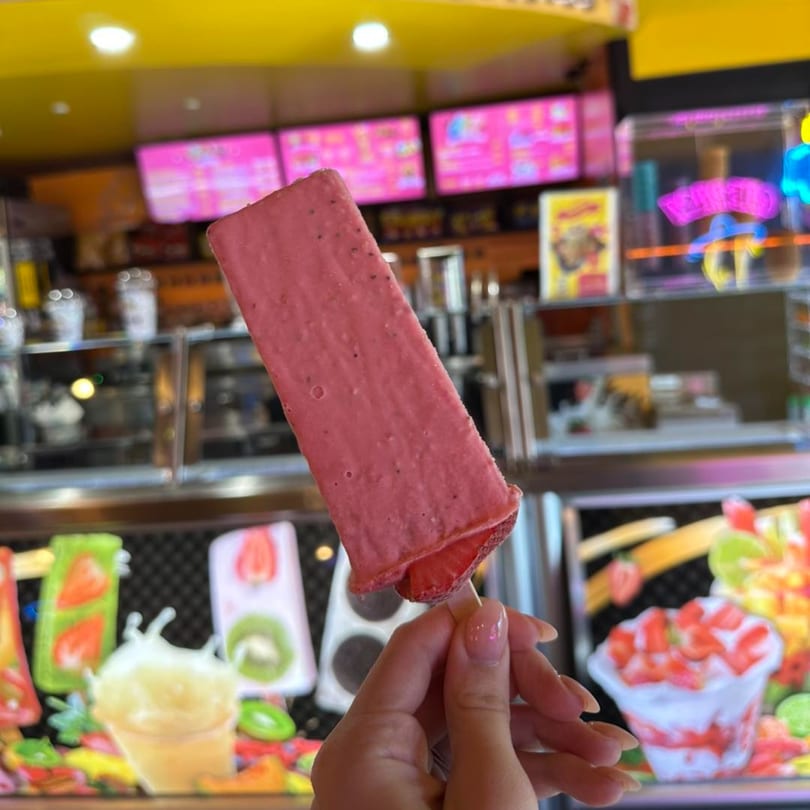Hand holding a pink strawberry paleta from Que Pasa Michoacana, with colorful ice cream displays in the background.