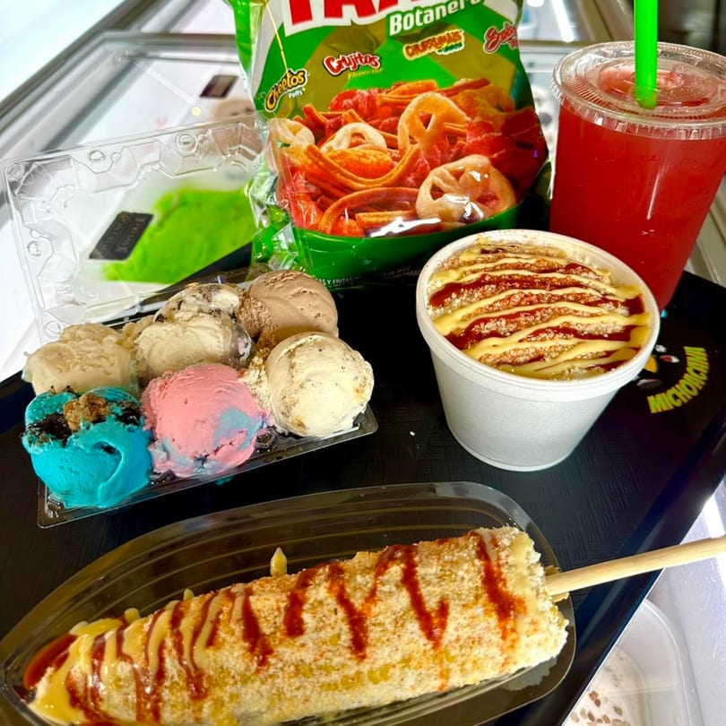 A tray of colorful ice cream scoops, elote, a red drink, and a bag of chips.